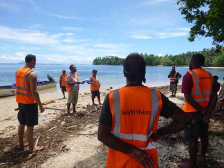 Aid workers in orange vests stand on a beach