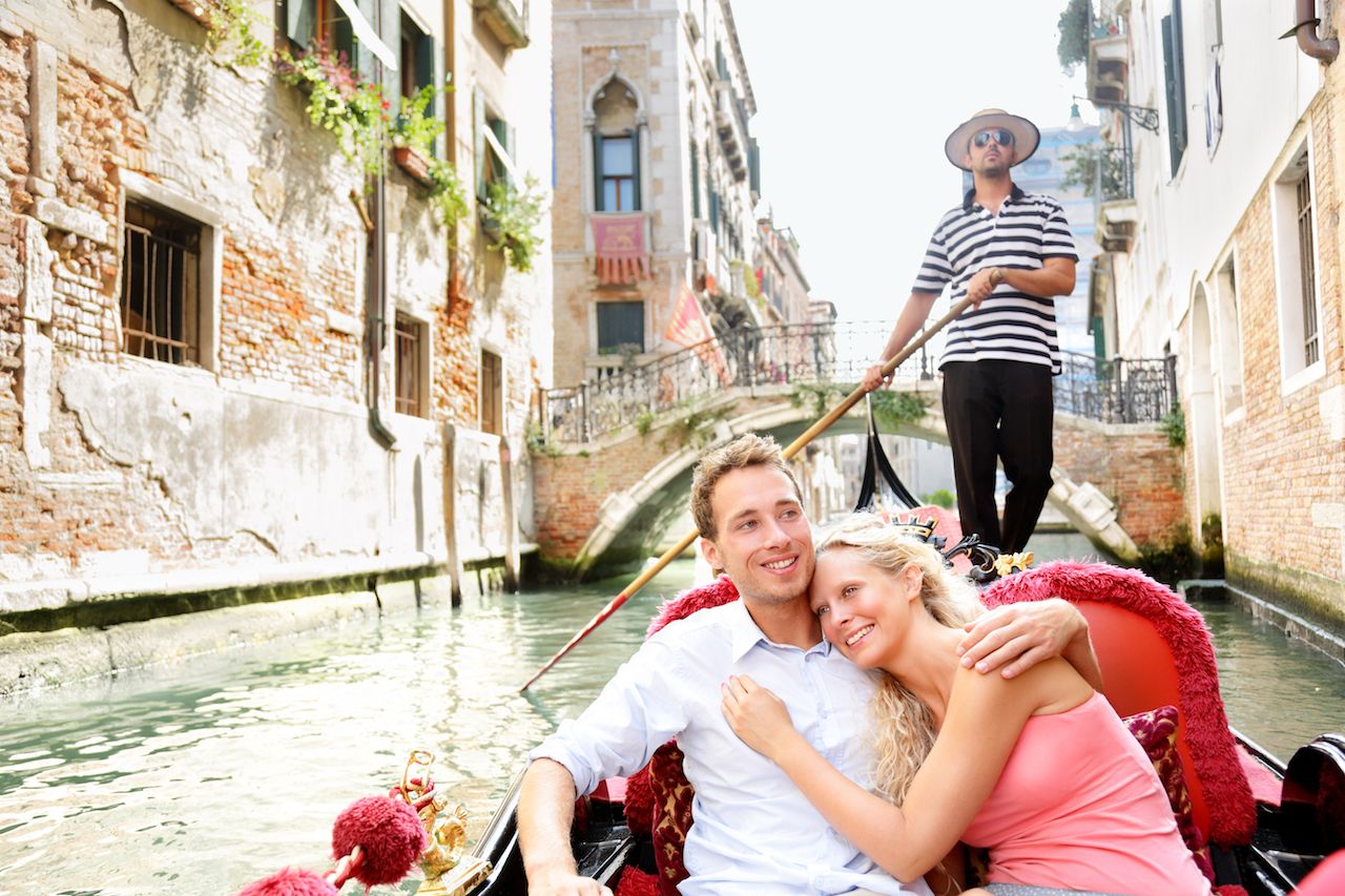 Romantic travel couple in Venice on Gondole ride romance in boat happy together on travel vacation holidays. Romantic young beautiful couple sailing in venetian canal in gondola. Italy, Europe.