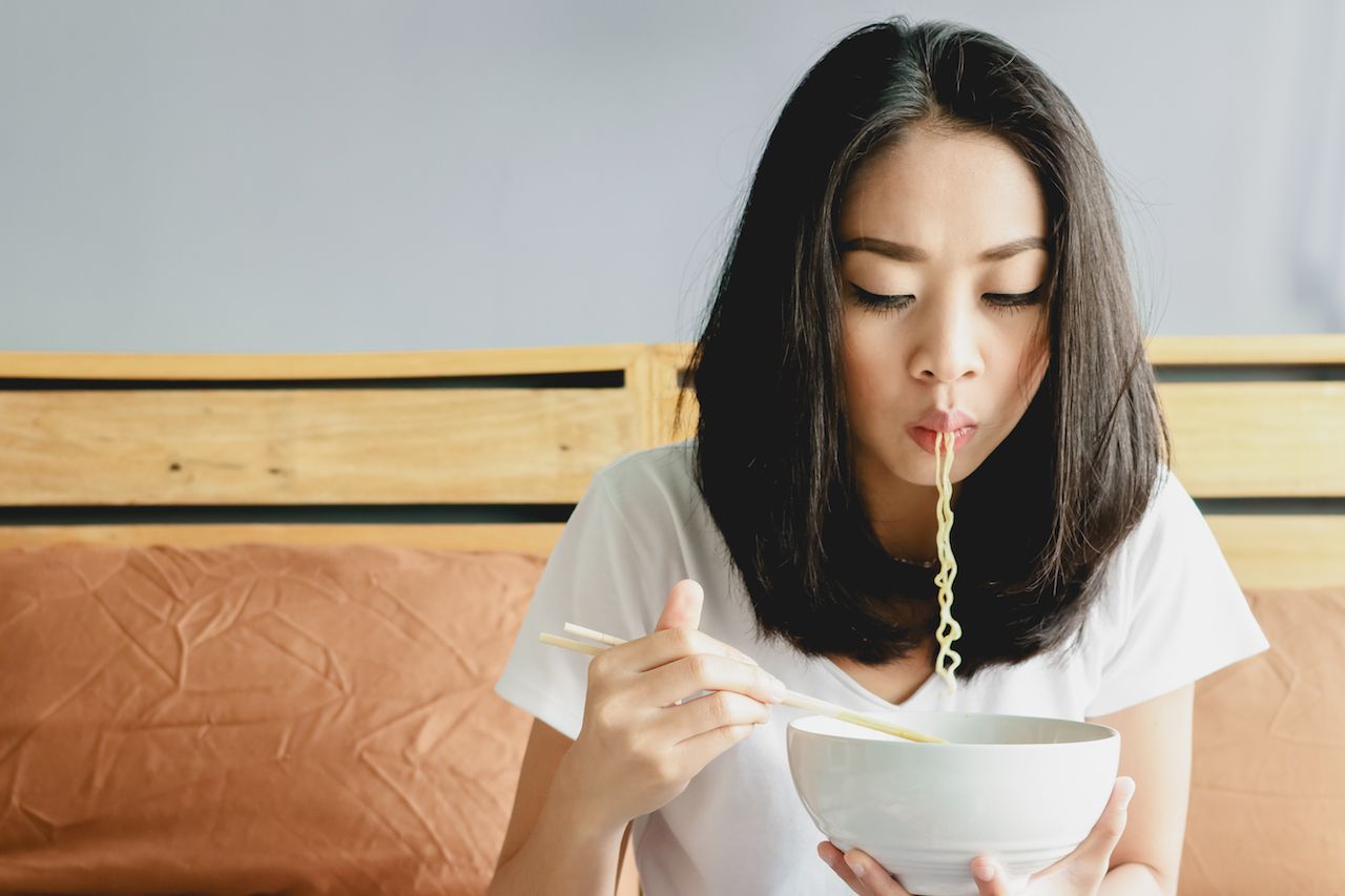 Asian girl eats instant noodle on the bed and noodle still in her mouth