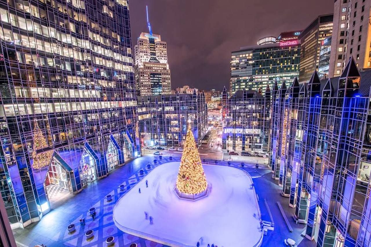 ice skating rink surrounding a christmas tree lit up under an illuminated city skyline at night