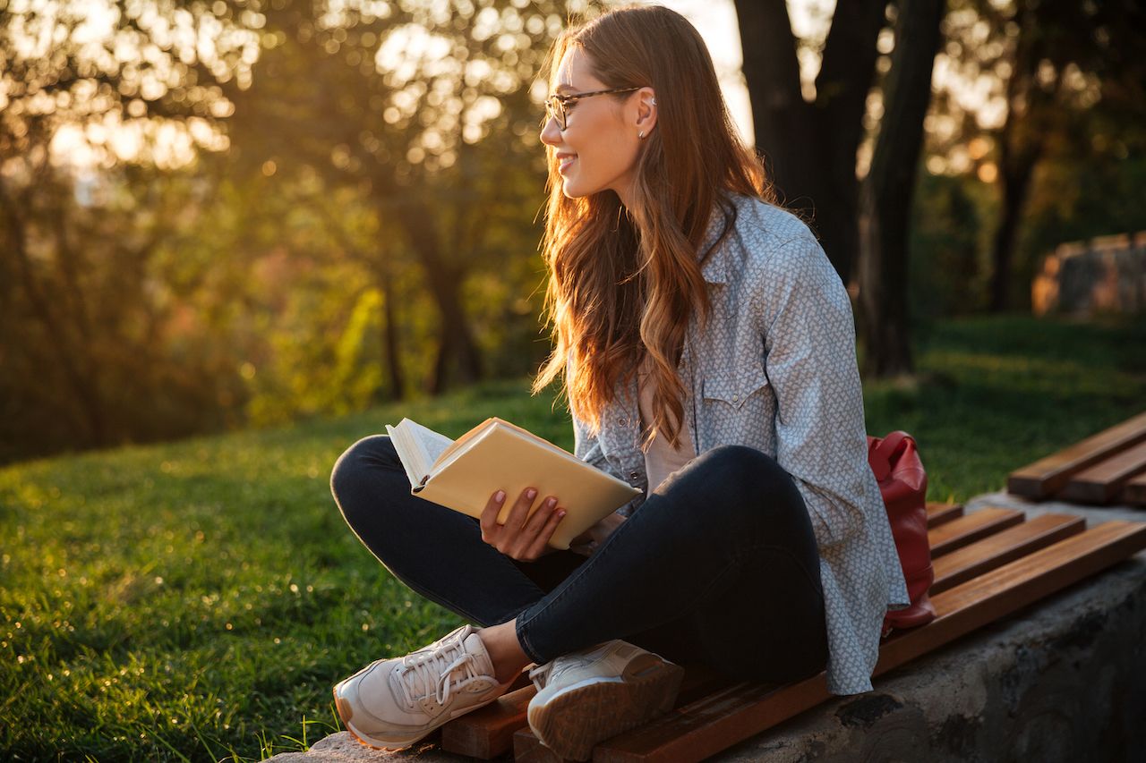 Side view of happy brunette woman in eyeglasses sitting on bench and holding book while looking away in park