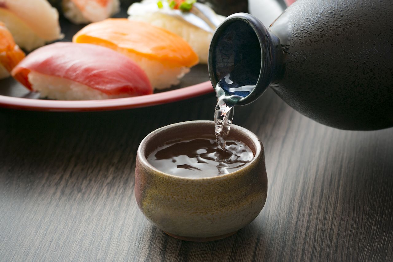 pouring Japanese sake from a black carafe into a cup with a platter of sushi in the background