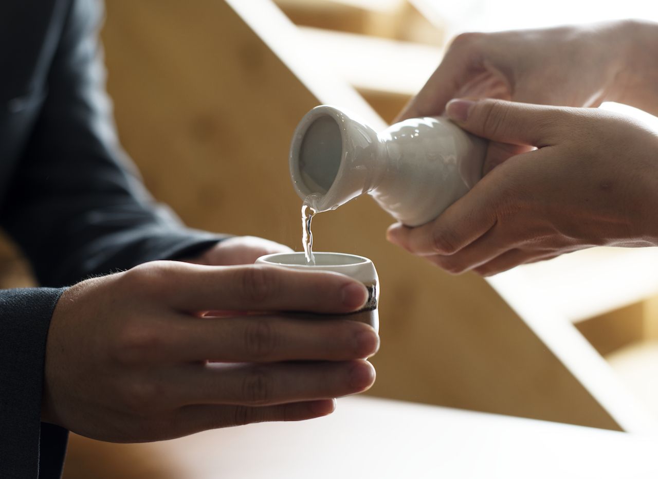 Person pouring sake from a white carafe with two hands into a white cup another person is holding