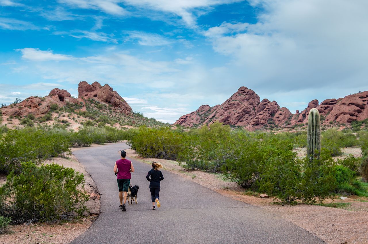 A couple and their dog jog down a trail in Phoenix, Arizona toward the iconic red sandstone buttes of Papago Park