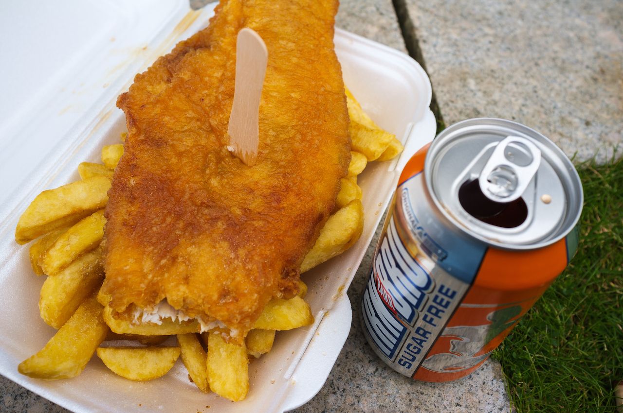 Fish and chips in a Styrofoam to-go container on the concrete beside a can of soda-Scottish foods