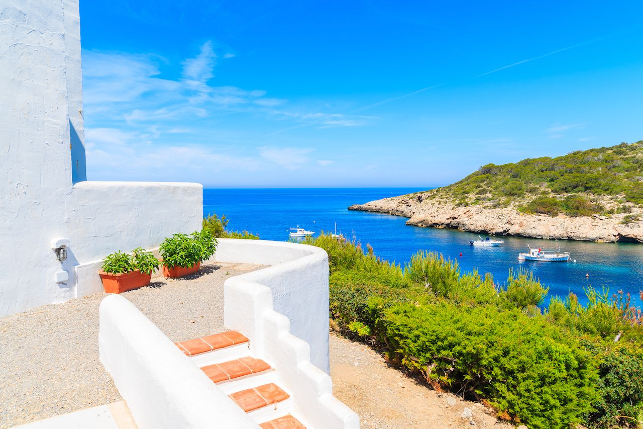 Steps to traditional white house and view of fishing boats on sea in Cala Portinatx bay, Ibiza island, Spain